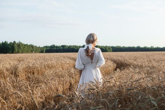 A blonde woman in a long white dress walks in a wheat field. The concept of a wedding and walking in nature.