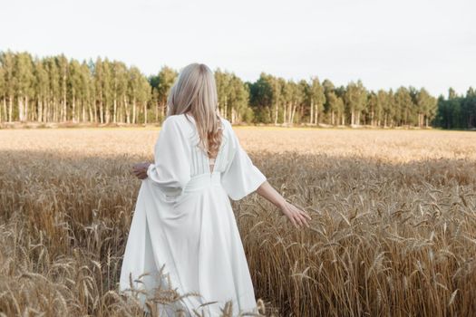 A blonde woman in a long white dress walks in a wheat field. The concept of a wedding and walking in nature.