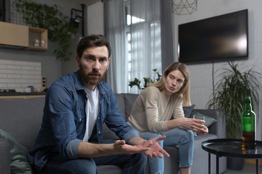 Female alcoholism, husband in despair, woman wife drinking hard alcohol, couple sitting on sofa in living room