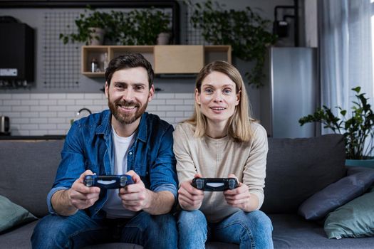 Happy couple man and woman playing game using joystick while sitting on sofa at home
