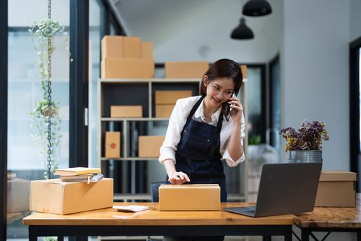 Business owner talking on mobile phone and taking order. Female asian entrepreneur working at home office confirming the order on phone