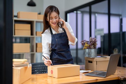 Business owner talking on mobile phone and taking order. Female asian entrepreneur working at home office confirming the order on phone