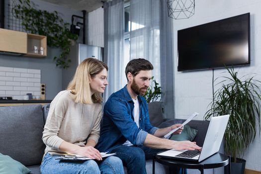 Young family, husband and wife, reviewing their bills, loan and mortgage agreements, sitting on the couch at home, working with documents on a laptop, embarrassed, upset