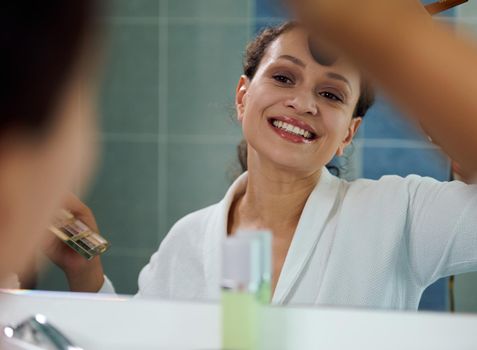 Pleasant stunning woman, in white bathrobe, reflecting in the mirror, smiles a beautiful toothy smile while applying a mattifying powder on her face, satisfied of her appearance and natural make-up