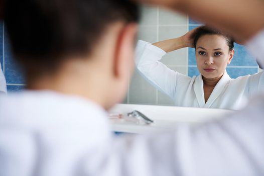 Soft focus on a beautiful dark-haired middle-aged woman in a white bathrobe, reflecting in the mirror, tying a ponytail during daily morning routines in the bathroom. Beauty advertisement. Body care