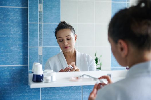 Beauty portrait of a reflecting in the bathroom mirror, attractive young woman in her 30s wearing a white waffle bathrobe, squeezing out moisturizer and applying it to her hands. Skin and body care