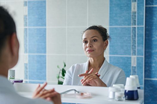 Beautiful middle-aged Hispanic woman, wearing a white waffle bathrobe, looking at her reflection in the bathroom mirror. Daily morning beauty routines. Home spa. Natural beauty people. Aging process