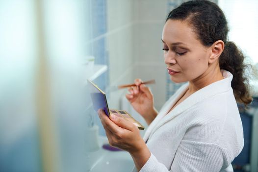 Side view. Close-up of a stunning pretty woman, wearing a white bathrobe, holding a make-up brush and a fashionable colorful eyeshadows palette, applaying natural make-up, standing in the bathroom