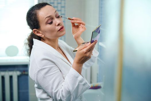 Beautiful middle-aged Hispanic woman in white bathrobe, holding a colorful eyeshadow palette and looking at her reflection in mirror while applying makeup on upper eyelids. Beauty and Fashion concept