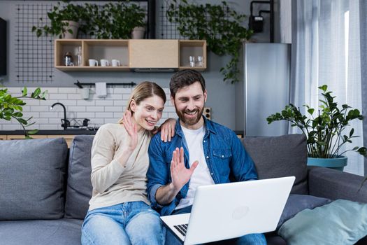 Young couple, family, man and woman sitting at home on the couch talking on video call from laptop to family, friends. They wave, smile, greet