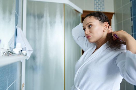 Middle-aged pretty woman, wearing white waffle bathrobe brushing her beautiful hair with comb while looking at her reflection in mirror in bathroom interior. Daily morning routines, hair care concept.