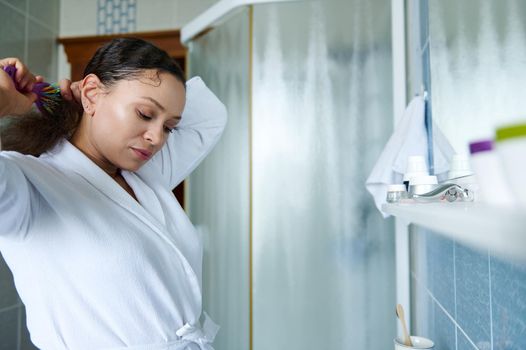 Multiethnic dark haired pretty woman brushing her curly hair with comb, standing in front of of a mirror in a home bathroom interior. Daily morning routines, hair care concept.