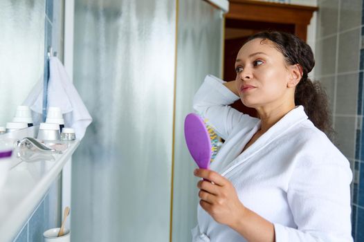 Portrait of a beautiful dark-haired middle-aged woman in a white bathrobe, combing hair, caring for curly chevelure, during morning routines, in the bathroom. Hair care, home spa, beauty treatment