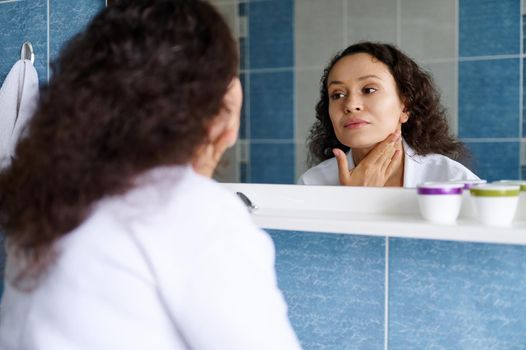 Charming multiracial 35s young brunette, beautiful woman in a white bathrobe examines her skin in the mirror, for the first signs of aging and wrinkles, applying moisturizer and massaging her face.