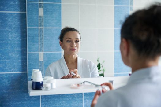 Gorgeous middle-aged African American woman in a white waffle bathrobe looks at her reflection in the bathroom mirror while applying moisturizing cream on her hands. Skin and body care concept