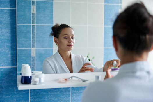 Charming middle aged dark-haired woman in a white bathrobe, reflected in the mirror, squeezing some moisturizer on her hands during daily beauty procedures in the bathroom. Skin and body care concept