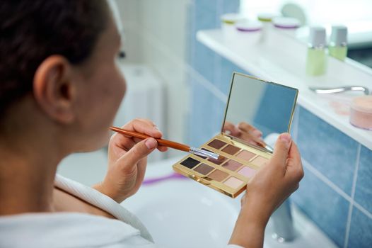 Rear view of a blurred dark-haired young woman, in a white bathrobe, holding eyeshadow palette with small cosmetic mirror and makeup brush, while applying make-up, standing in the home bathroom