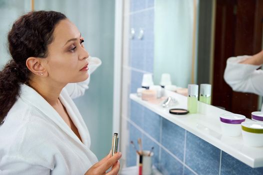 Charming African American woman wearing a white bathrobe, applying make-up, using mascara, augmenting the volume to her eyelashes, looking at her reflection in the bathroom mirror. Beauty fashion.