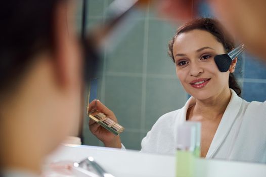 Reflecting in the mirror, delightful stunning young woman in a white waffle bathrobe, applies a mattifying powder on her face, satisfied of her appearance and natural make-up. Natural beauty people