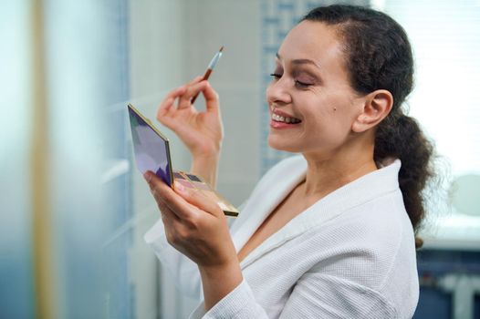 Attractive stunning middle-aged curly haired woman, wearing a white bathrobe, holding a colorful makeup palette, cutely smiles while is applying eye shadows to upper eyelid, standing in the bathroom