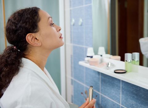 Side portrait of a charming woman in a white bathrobe, applying make-up, using mascara, augmenting the volume to her eyelashes, looking at her reflection in the bathroom mirror. Beauty fashion.