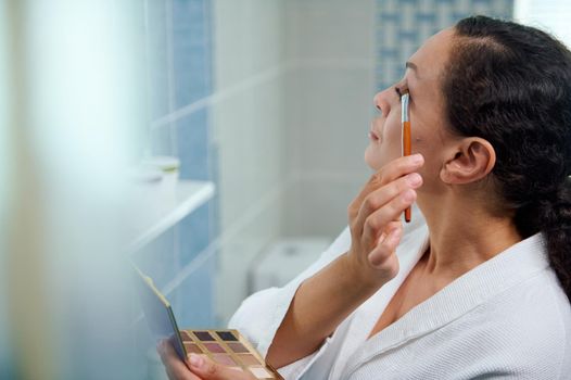Close-up. Stunning beautiful middle-aged multiethnic woman in a white bathrobe holds a colorful eyeshadow palette and using a make-up brush, applies eye shadow to upper eyelid. Beauty, make-up concept