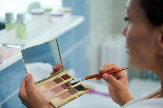 Soft focus on beautiful eyeshadow palette with small cosmetic mirror and makeup brush in hands of blurred young woman applying make-up while getting ready, in home bathroom interior