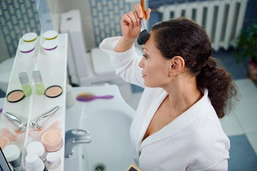 View from above of a delightful dark-haired beautiful woman in a white waffle bathrobe, applying a mattifying powder on her face, satisfied of her appearance and natural make-up. Natural beauty people