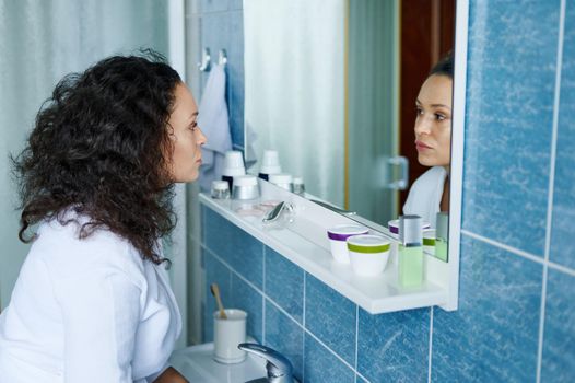 Close-up of a multiethnic attractive 35s woman in white bathrobe standing in bathroom reflected in mirror, thinking about her appearance. Beauty morning routine, body, skin and hair care concept