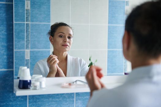 Reflection in the bathroom mirror of serene beautiful middle-aged woman massaging her hands soaked in moisturizer, caring for the skin of her hands.