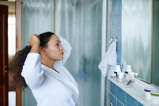 Dark haired beautiful multiethnic mature woman, wearing a white waffle bathrobe, combing hair, looking at her reflection in the bathroom mirror. Beauty and body care concept