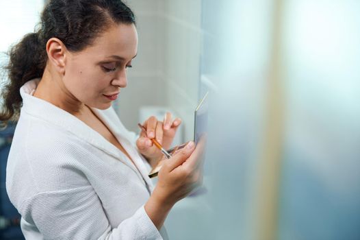 View through blurred transparent doors of a shower stall on a charming elegant brunette, middle-aged multiethnic woman holding a fashionable colorful palette with eyeshadow and a makeup brush in hands