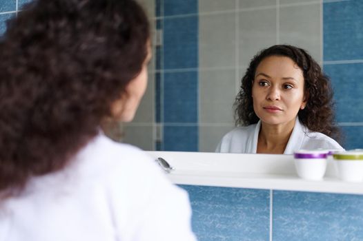 Beautiful curly brunette, Hispanic woman in a white bathrobe, reflecting in the mirror while admiring herself, examining the condition of the skin and wrinkles, standing at home in the bathroom