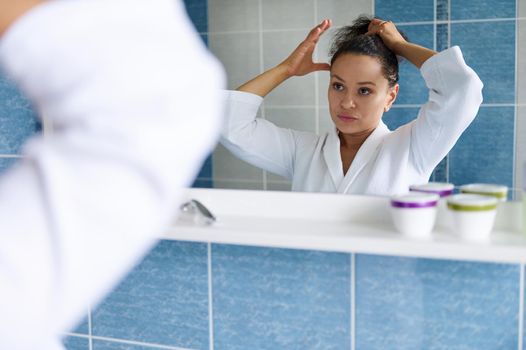 Beautiful dark-haired Hispanic woman in white bathrobe ties her ponytail and looks at her reflection in the bathroom mirror. Skin and body care. Daily home beauty and spa treatments. Morning routines.