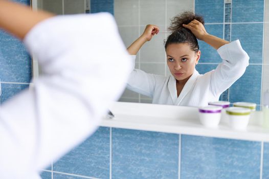 Beautiful multiethnic 35s young woman in white bathrobe ties her ponytail, looking at her reflection in the bathroom mirror. Skin and body care. Daily home beauty and spa treatments. Morning routines.