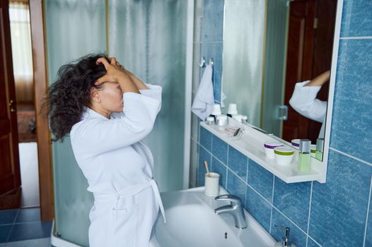 Close-up multiethnic attractive 35s woman in white bathrobe, standing in bathroom reflected in mirror, touching curly hair looking pensive thinks about hairstyle change. Beauty morning routine concept