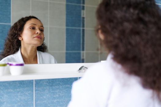 Middle-aged beautiful multi-ethnic woman in a white waffle bathrobe, admires herself in the mirror while performing morning beauty treatments in the bathroom. Skin and body care concept. Aging process