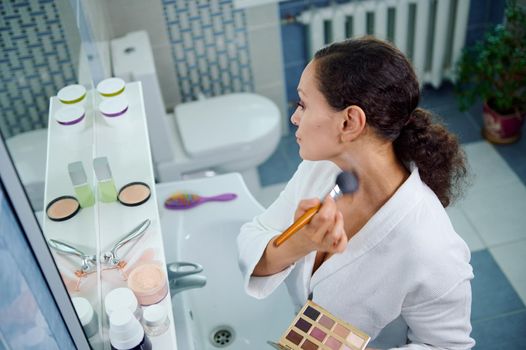 Top view of a stunning multiethnic woman, wearing a white bathrobe holds a colorful makeup palette and applies mattifying powder on her face and neck, looking at her reflection in the bathroom mirror