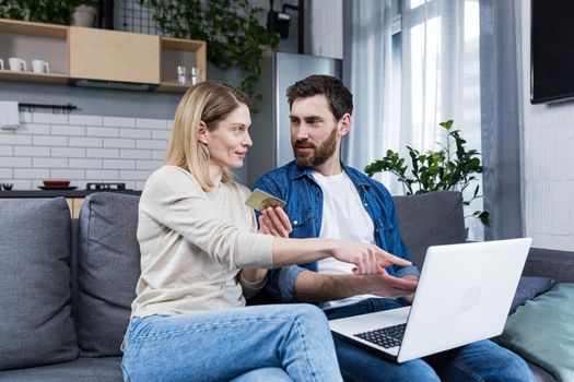 Happy married couple man and woman sitting at home on the couch using laptop for online shopping, holding a credit bank card