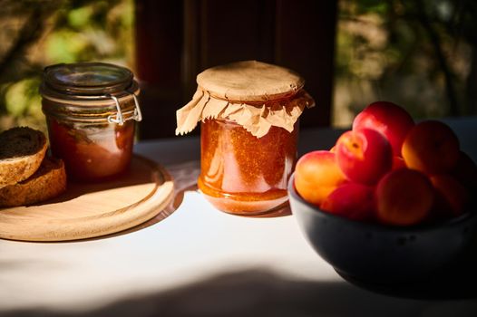 Still life composition of glass jars with delicious homemade jam, ripe red apricots in a blue ceramic bowl, on a table, against the background of wooden window, overlooking garden in rustic kitchen