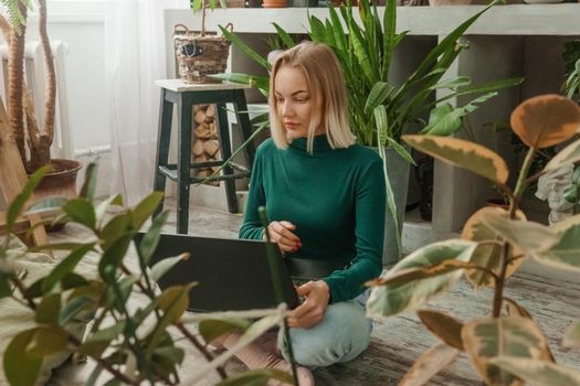 A blonde woman in a room with a lot of green indoor plants is working on a laptop. The concept of biophysical design in the interior. Work from home, work as a freelancer.