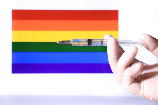 Hand in surgical glove holding syringe and rainbow flag in the background