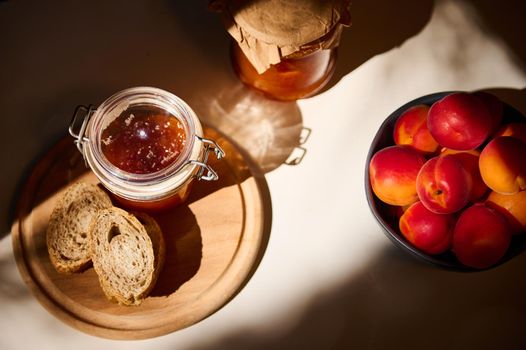 Top view. Still life composition of a homemade apricot jam and whole grain bread on a wooden board, ripe red apricots in a blue ceramic bowl on a table. Sunlight fall on the table