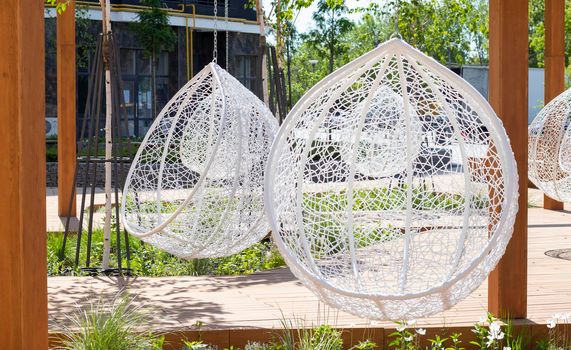 A cozy seating area in the yard of a modern residential complex with wooden benches, hanging white wicker rattan chairs, garlands and a beautiful landscape design with flowers and a lawn
