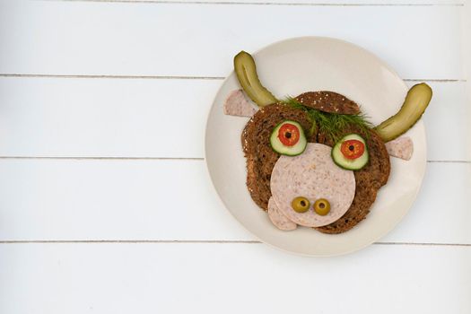 Sandwich in the form of a portrait of a cheerful bull made of dark bread, sausages and vegetables on a plate, on a white wooden background, copispice. High quality photo