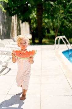 Little girl eating a piece of watermelon while walking by the pool with turquoise water. High quality photo