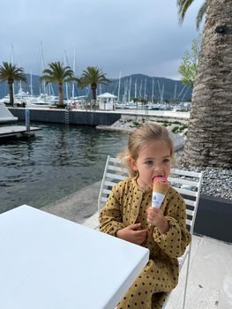 Little girl sits at a table on the embankment and eats an ice cream cone. High quality photo