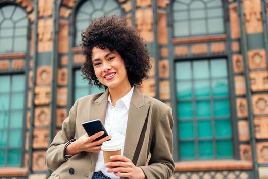 young business woman smiles happy looking at camera with a mobile phone in one hand and a coffee in the other, concept of urban lifestyle and technology, copy space for text