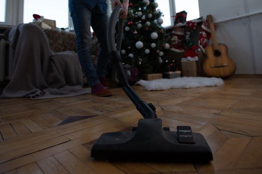 Young woman cleaning with vacuum cleaner, vacuuming under Christmas Tree needles with New Years ornaments on hardwood wooden floor