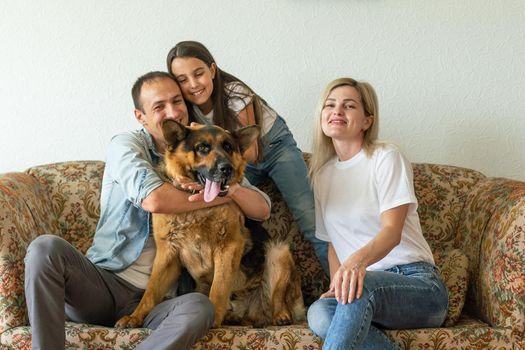 Beautiful young parents, their cute little daughter looking at camera and smiling, sitting with their cute dog on sofa at home.